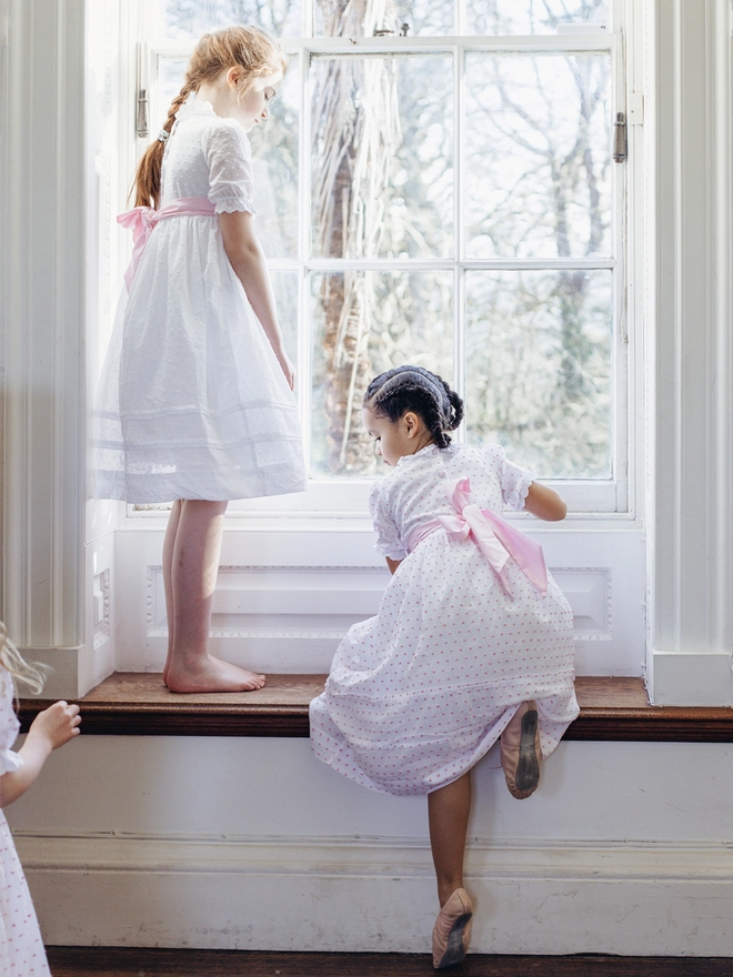Two girls climb on a window sill in white dresses with pink sashes