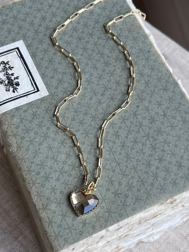 Gold paperclip chain necklace with smokey grey heart charm on a pale green book