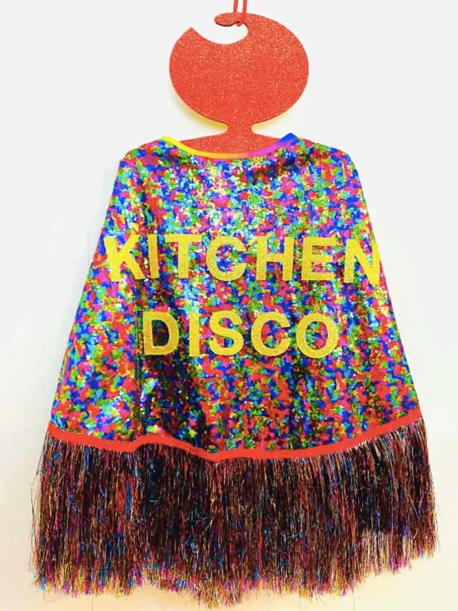 A multi-coloured maxi length cape with 'KITCHEN DISCO' across the back in gold text. The tinsel is rainbow,