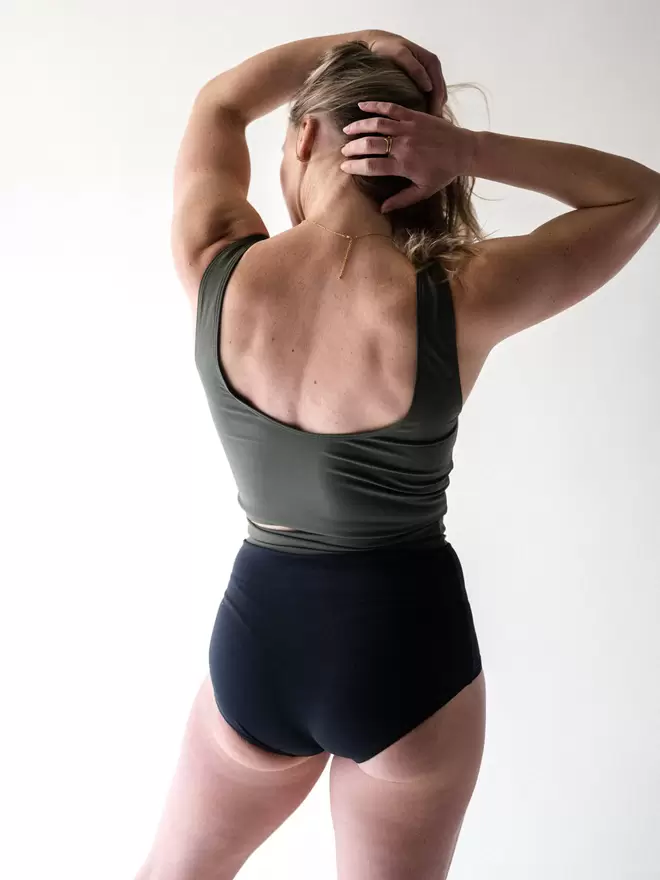 Back view of woman with hands in hair wearing Davy J Sustainable Waterwear olive green crop swim top with scoop back and black high waist bikini briefs rolled down to show olive green lining