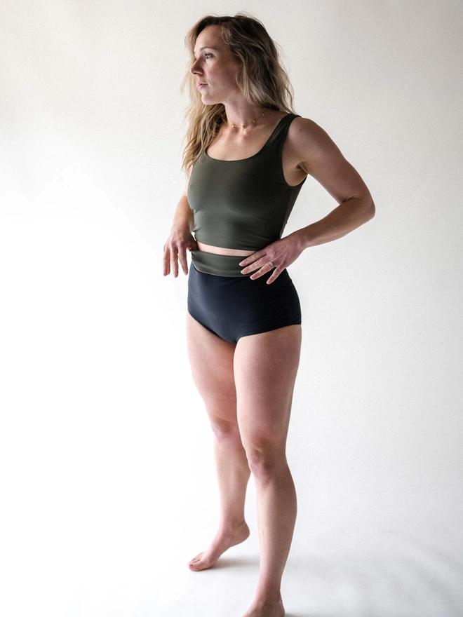 A lady with blonde hair stood on tip toes, wearing a Davy J Sustainable Waterwear olive cropped swim top with squared neckline and shoulder straps. Worn with a pair of black high waisted bikini briefs with the waist rolled down showing a band of olive lining  Edit alt text