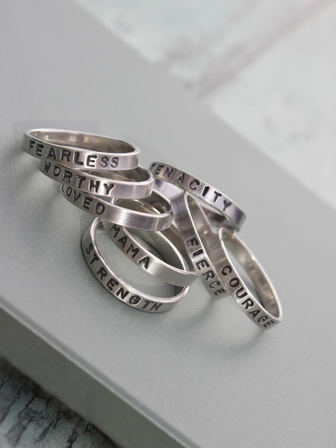 A selection of sterling silver plain band rings with a single stamped word, arranged in a casual pile on top of a green ceramic tile on a green background.