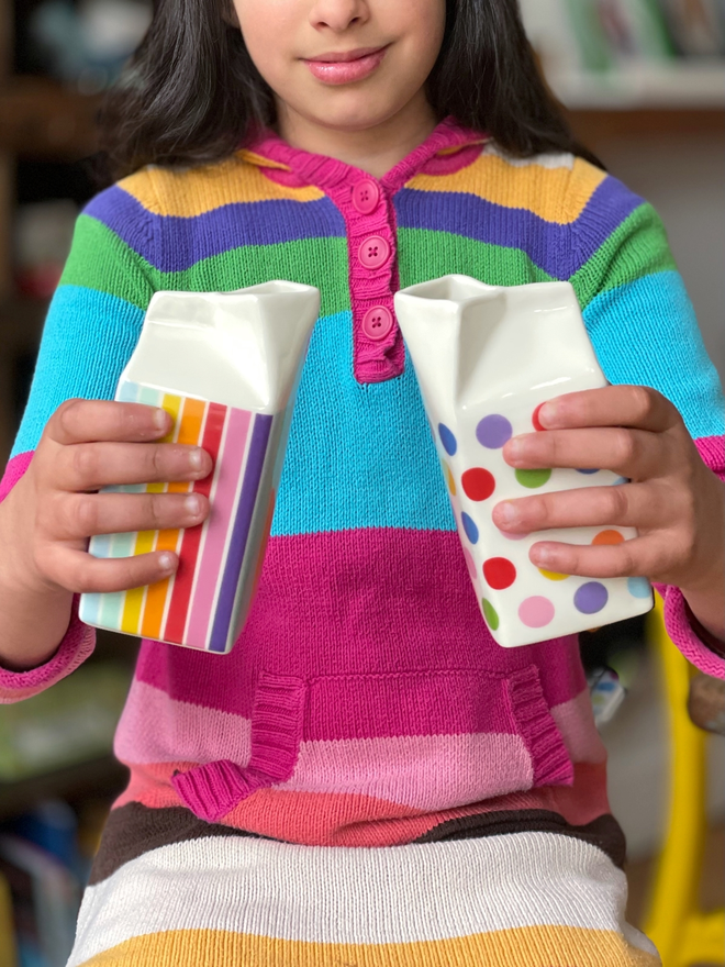 Smiling child in striped jumper holding two porcelain milk carton jugs - one striped, one polka dot - in red, orange, yellow, green, blue, purple, and pink colours