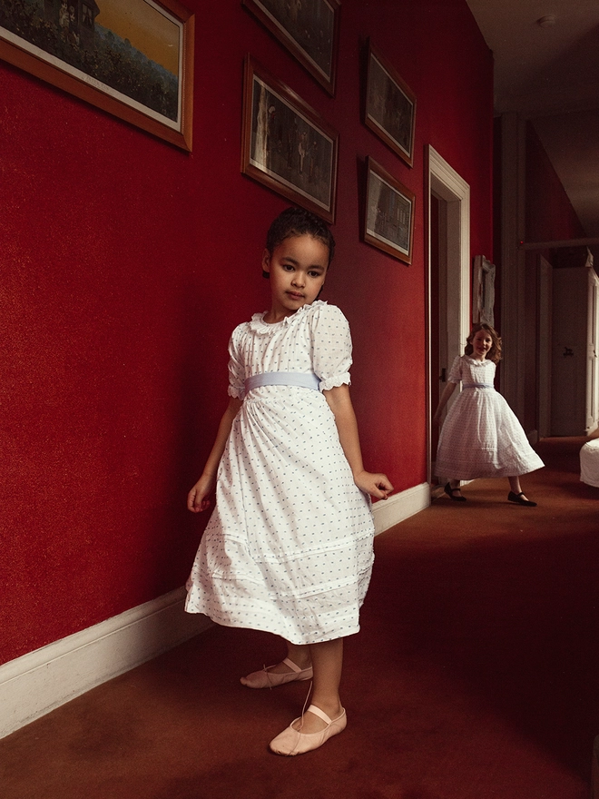 A little girl does a twirl in a red corridor in a blue and white dress with a girl stepping out of a doorway behind