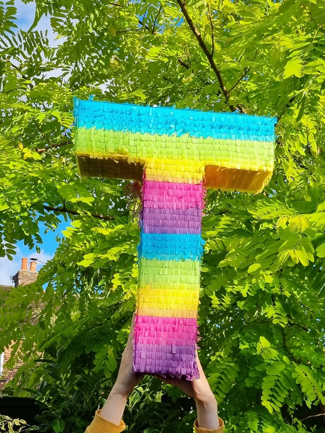 rainbow stripe letter t pinata by pinyatay held up in the sunshine