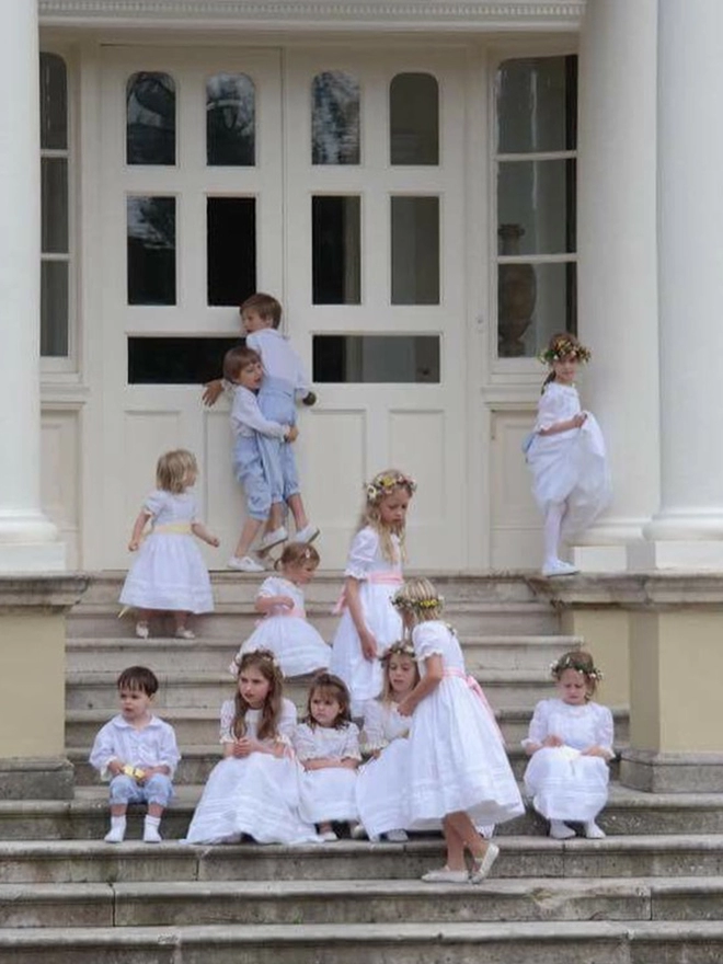 A group of children in white dresses with pink sashes sit on the steps of a big house