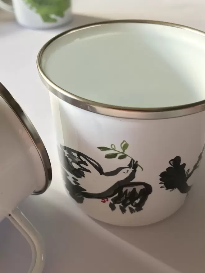 Close up of a white enamel mug decorated with a black peace dove design