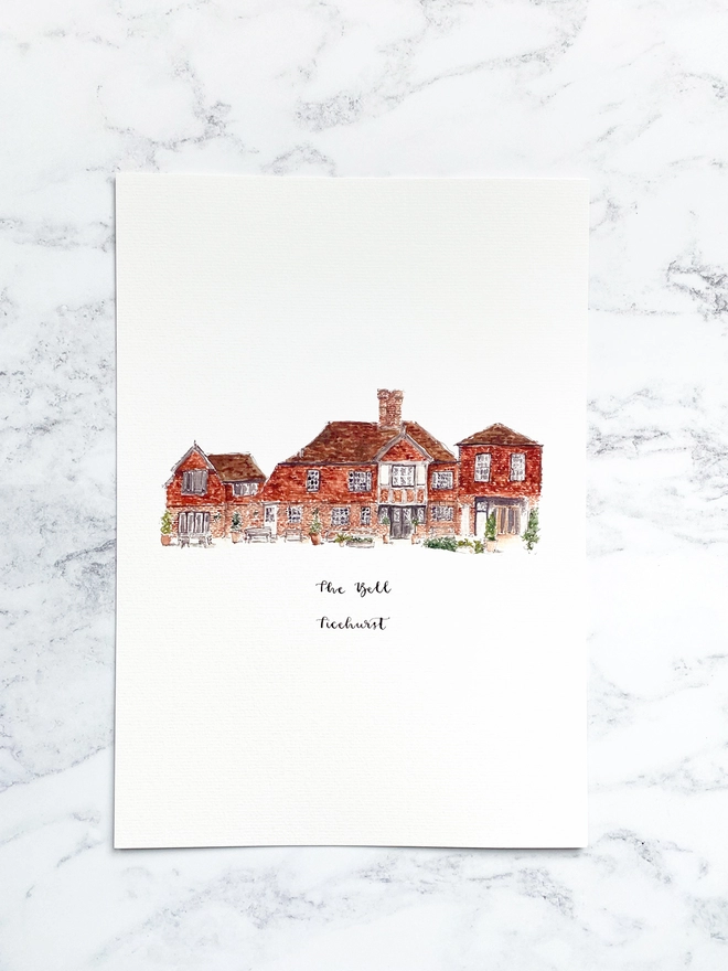 Watercolour painting of The Bell in Ticehurst, a beautiful brick building with a teracotta tile roof, small black framed white windows and seats and planting outside. The watercolour style is painted with a black pen outline and organic loose style with small details. The print is a small illustration on the centre of a white page and the paper sits on a white marble background.