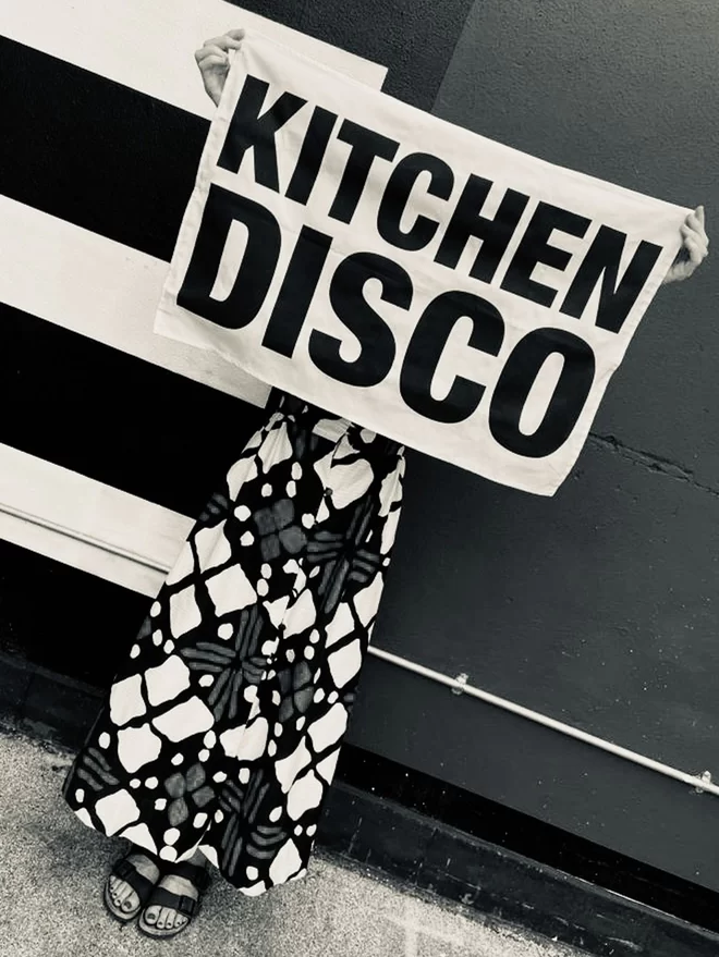 London Drying Kitchen Disco black screen printed text on white tea towel being held up by person in a patterned dress in front of a half solid colour/half striped painted wall 