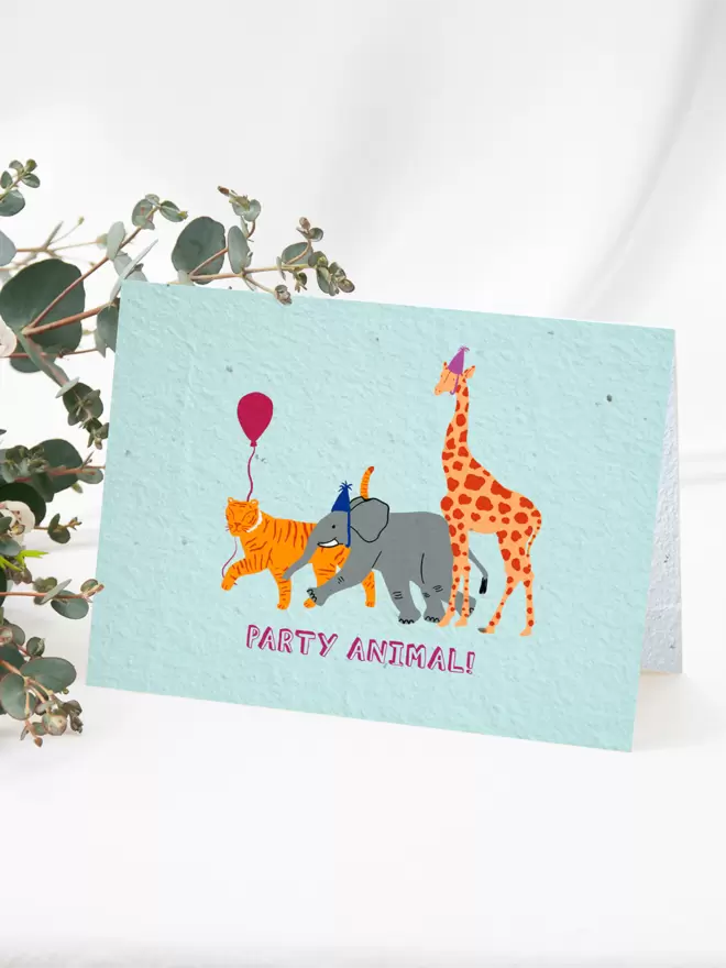 Plantable Card with illustration of an elephant and giraffe wearing party hats and a tiger with a balloon with ‘Party Animal’ written beneath on a blue background with some flowers next to the card in the background