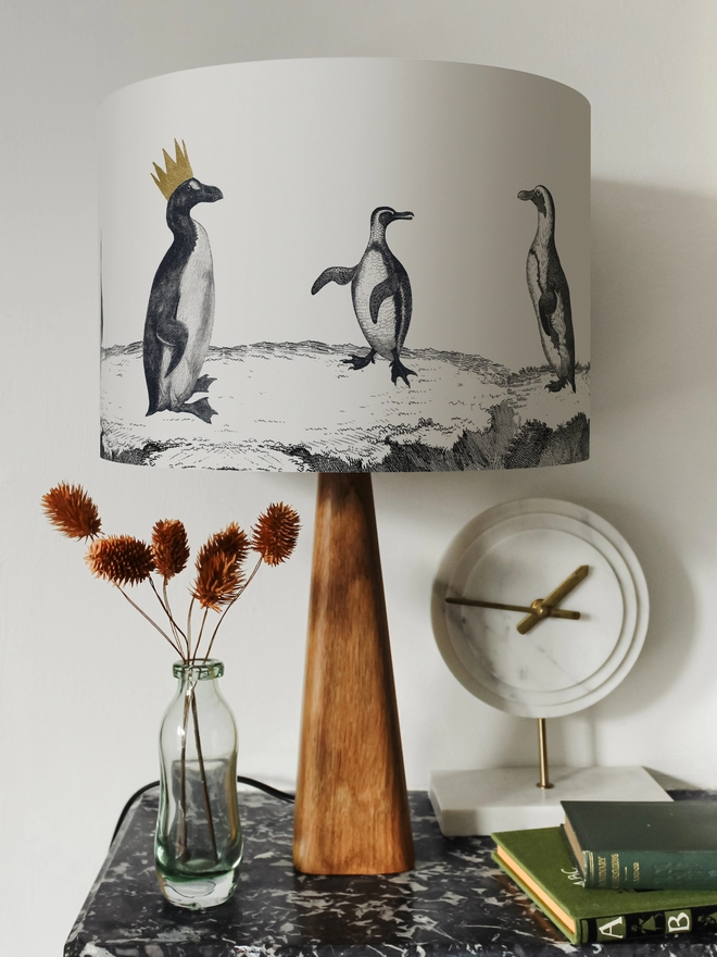 Drum Lampshade featuring a parade of penguins on a wooden base on a shelf with books and ornaments
