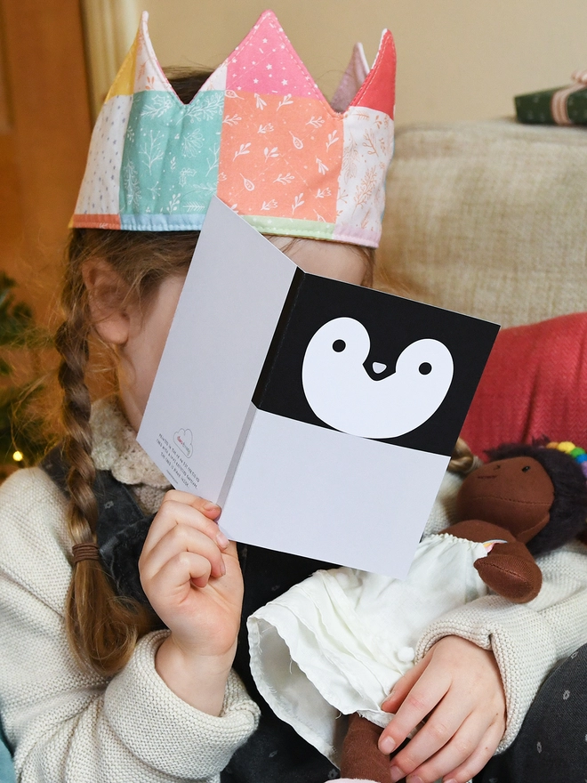 A young girl wearing a pastel patchwork crown holds a penguin greetings card in front of her face.