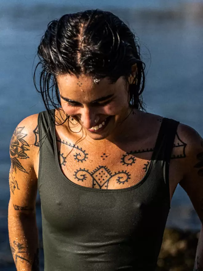 A lady with wet dark hair and lots of tattoos looking down at the floor smiling and wearing a Davy J Sustainable Waterwear olive cropped swim top with square neckline and shoulder straps