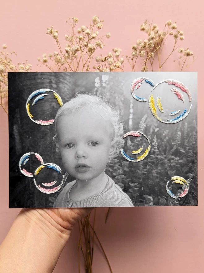 Black and white photograph of child with hand embroidered bubbles in shades of pink, yellow and blue