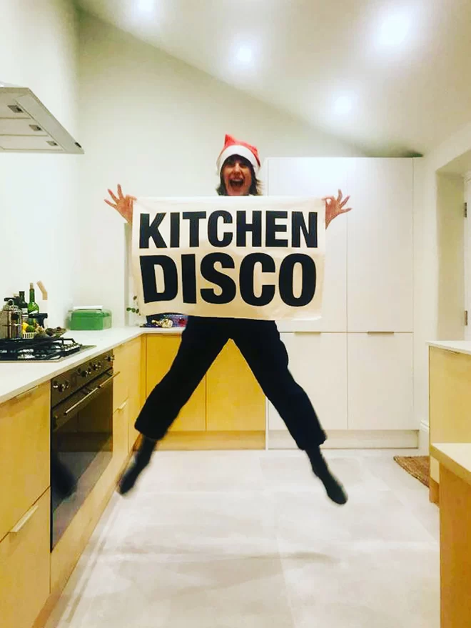 London Drying Kitchen Disco tea towel being held by person wearing a Santa hat jumping in a kitchen