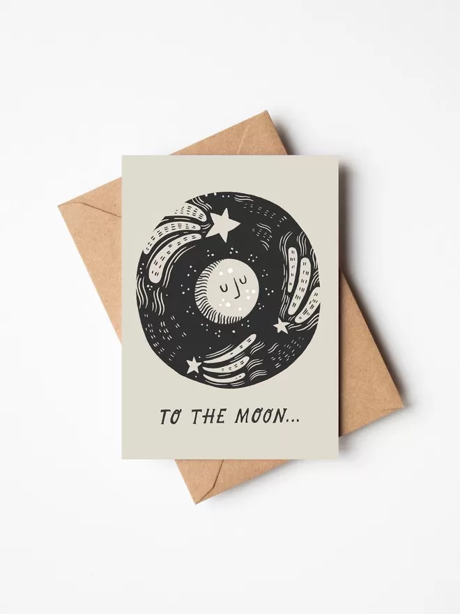 Black and white greeting card with illustration and the words to the moon written on it with a brown envelope underneath it