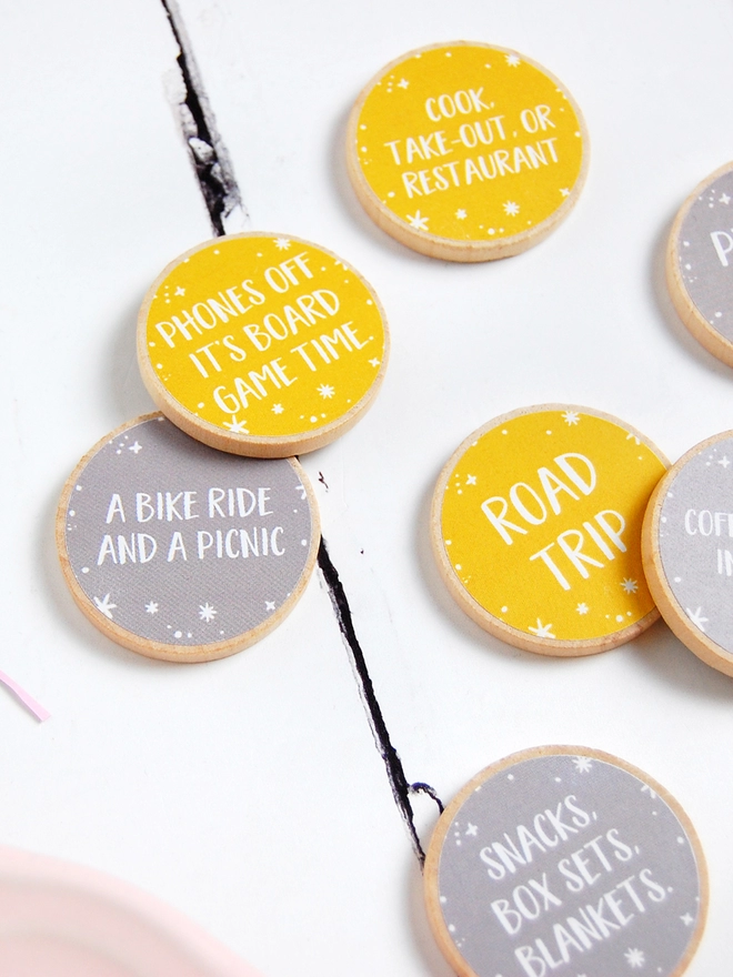 Several wooden tokens with yellow and grey labels, each with a friendship date idea on, lay on a white wooden surface.