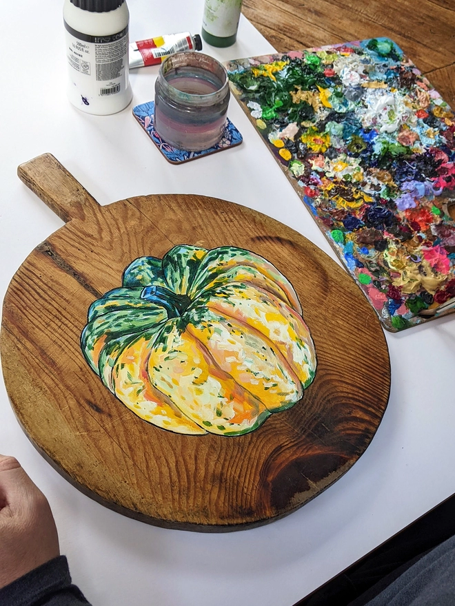 Wooden chopping board with carnival squash painted design on a table next to paints and painter's palette