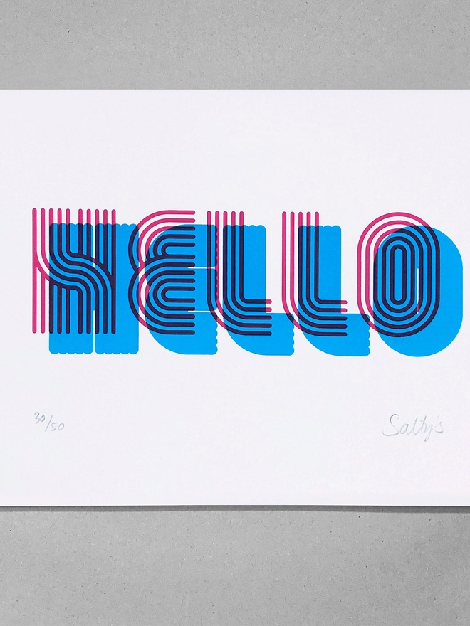 A flat lay of this print on a grey background. Stripy tram lines make up this font in a retro/sporty look. Hello is screenprinted in this font in magenta, with a cyan solid shadow beneath. 
