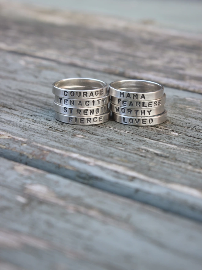 A collection of sterling silver simple plain band rings with words of empowerment stamped on them, stacked together in two towers side by side on a weathered blue painted table.