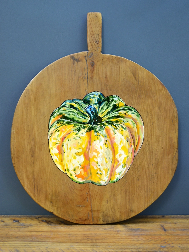 Wooden chopping board with handpainted design of a carnival squash standing against a wall