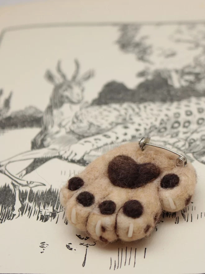 The underside of a needle-felted lucky leopard brooch, showing brown pads, resting on a vintage natural history book
