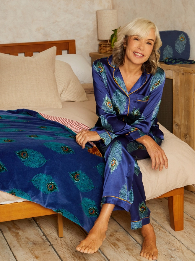 smiling blone sits on the end of bed with beige cotton bedding and velvet peacock feather throw wearing satin navy peacock feather print button up pyjamas.