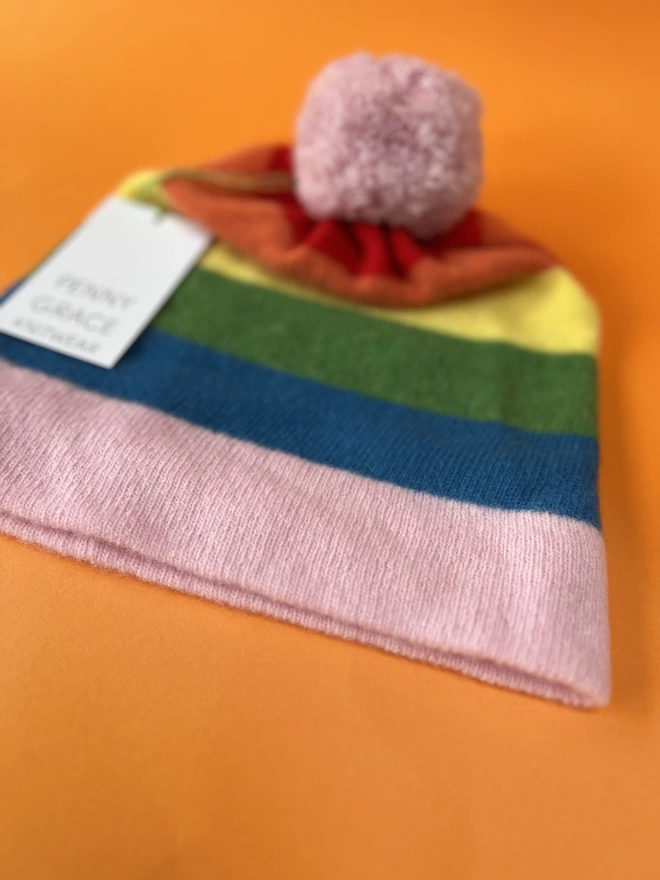 A close up of rainbow striped beanie hat shown at an angle on an orange background