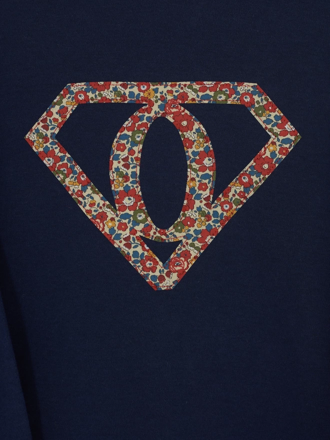 A navy long sleeve cotton t-shirt featuring the initial O inside a superhero motif. The motif is appliquéd in floral Liberty Print. A close up of the stitching.