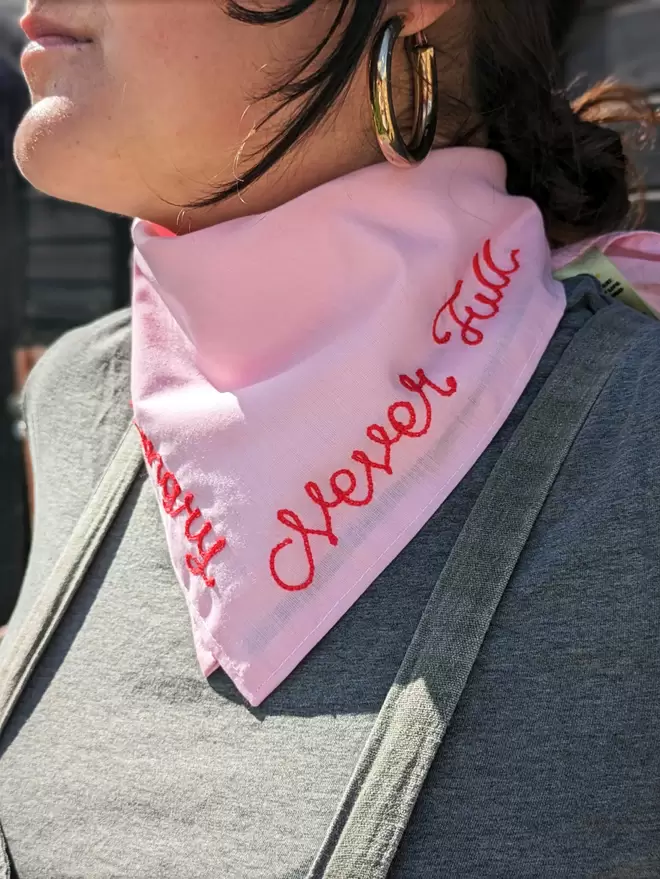 Woman wearing a pink Bandana Neckerchief with White Embroidered lettering readiing "Always Hungry, Never Full" in red embroidery thread