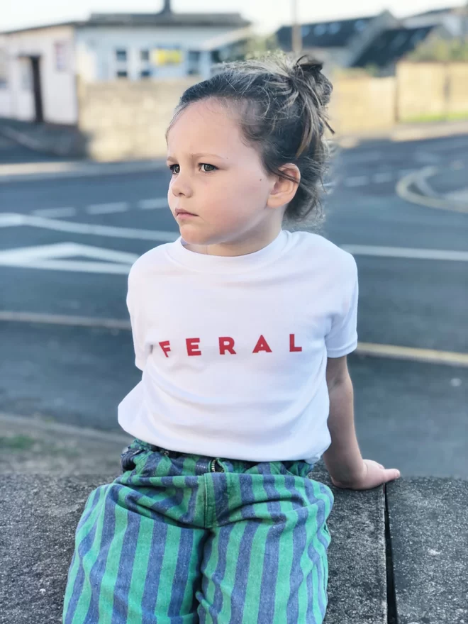 Photograph of a girl sitting on a wall wearing a white tshirt with the word FERAL screen printed on it in red ink.