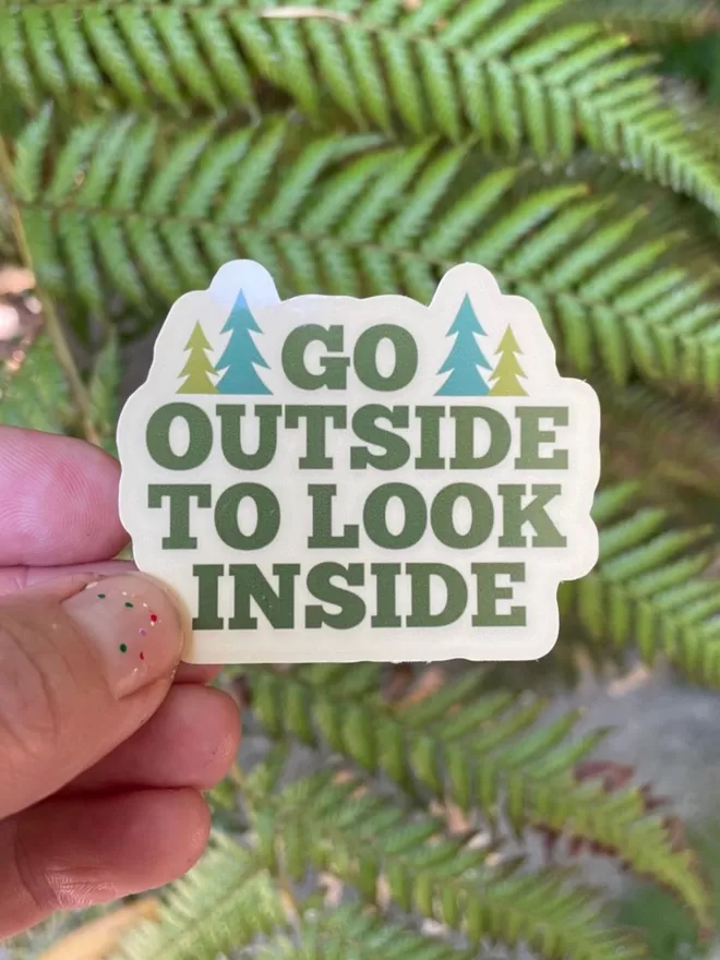 Go Outside To look Inside vinyl sticker being held in front of plants.