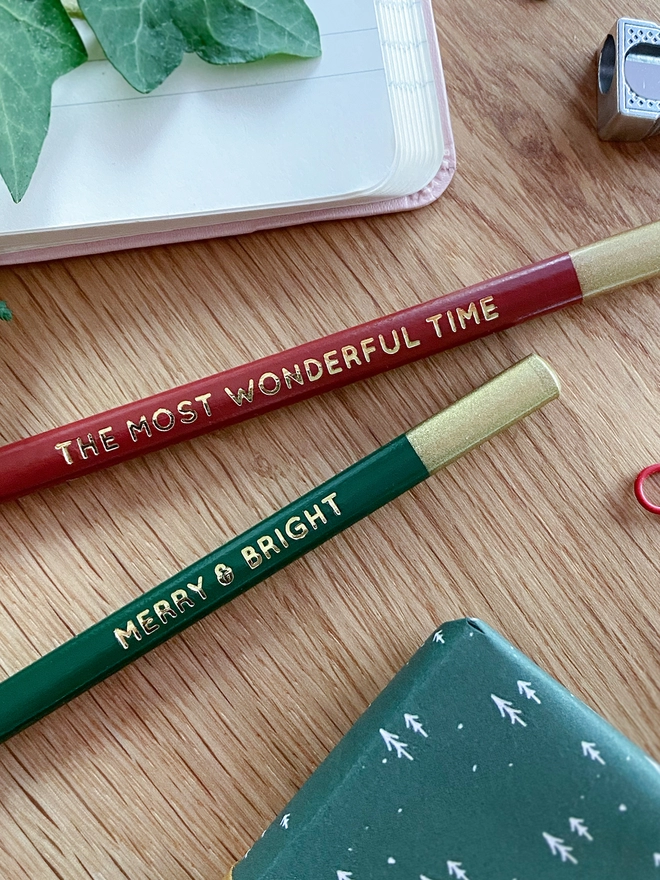 Two pencils, one red and one green, both with gold writing along the sides lay on a wooden desk.