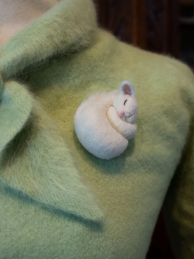 A needle felted white sleeping cat brooch on a green fluffy jumper 
