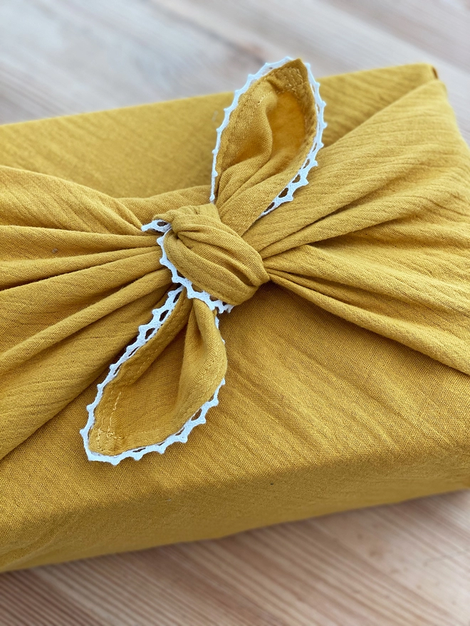 A gift wrapped in mustard yellow cotton fabric wrap with an ivory lace trim is on a wooden desk.