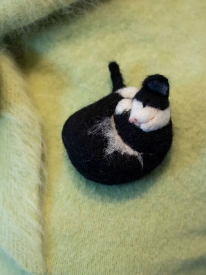 A needle feltedblack and white sleeping cat brooch on a green fluffy jumper 