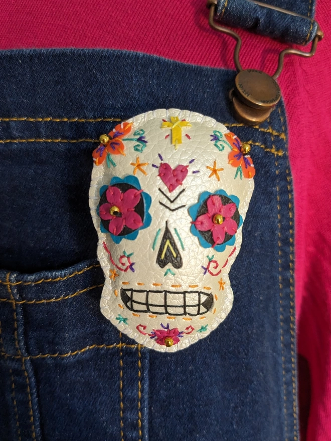 A hand stitched faux leather white Mexican Day of the Dead inspired sugar skull brooch with  colourful stitching and gold bead details, on a wooden background with red roses