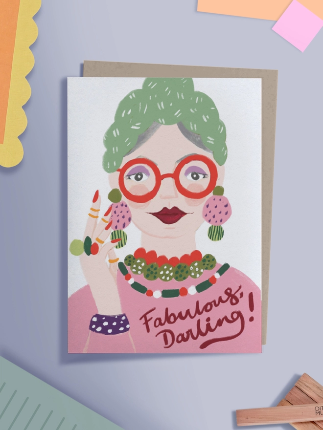 A flat lay of a greeting card that reads 'Fabulous Darling!' with an illustration of a glamorous older woman, wearing bold red glasses, and lipstick, with lots of colourful jewellery.