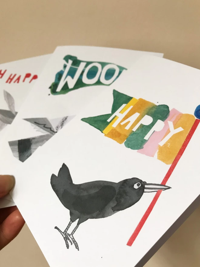 Set of three illustrated greetings cards by Esther Kent showing colourful lettering and stylised birds. The front card that reads 'HAPPY' on a flag help by a black painted bird.