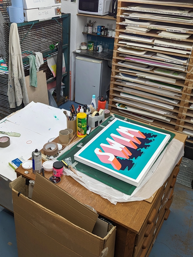 A pile of screenprints in a studio of the word SWIM in 3d typography in pink, orange and blue on a green background by artist Survival Techniques. The letters have a watery shadow.