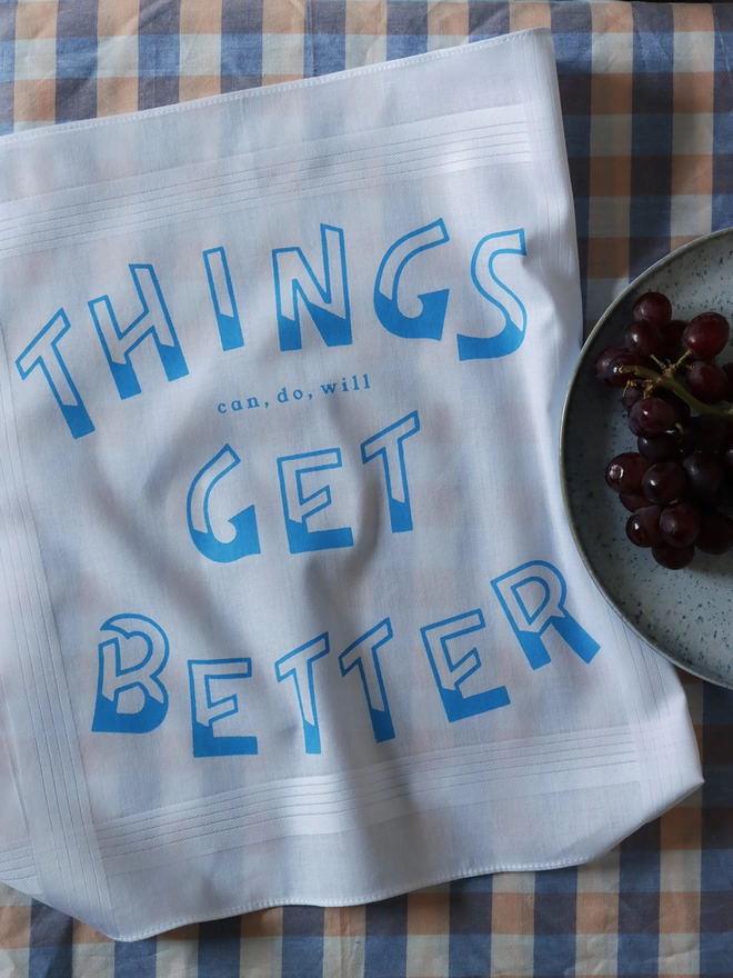 Mr.PS Things Get Better Hankie laid on a gingham tablecloth with a plate of grapes