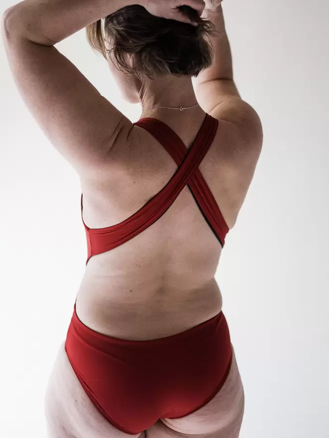 Woman in studio facing away with hands in her short brown hair, wearing red Davy J Sustainable Waterwear cutout swimsuit with wide cross back straps