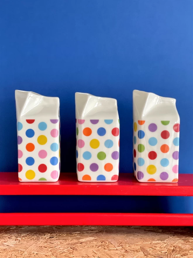 3 milk carton jugs red shelf , surface decoration is multicoloured polkadot pattern in red , blue , green , orange , purple and yellow ., the background is cobalt blue