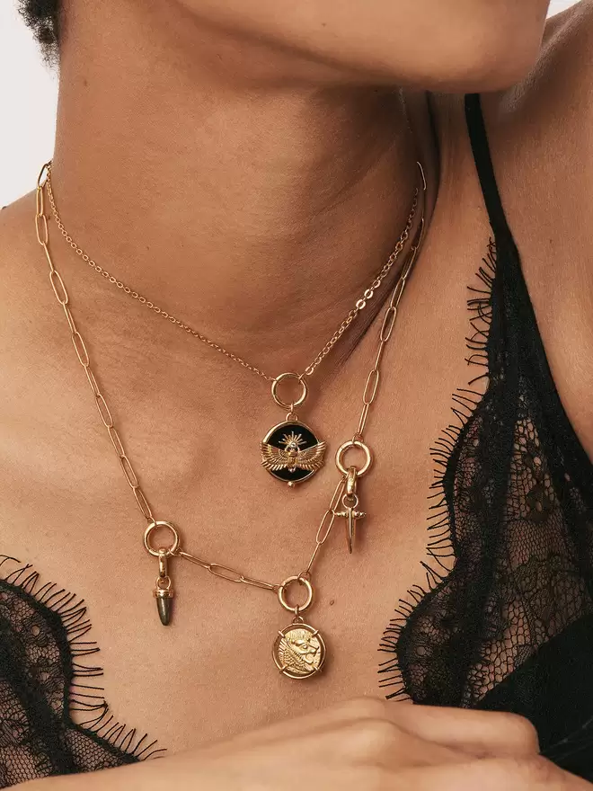 woman wearing two gold necklaces styled with a scarab beetle medallion, a hematite bullet charm, a lion coin medallion and a small bayonet dagger charm