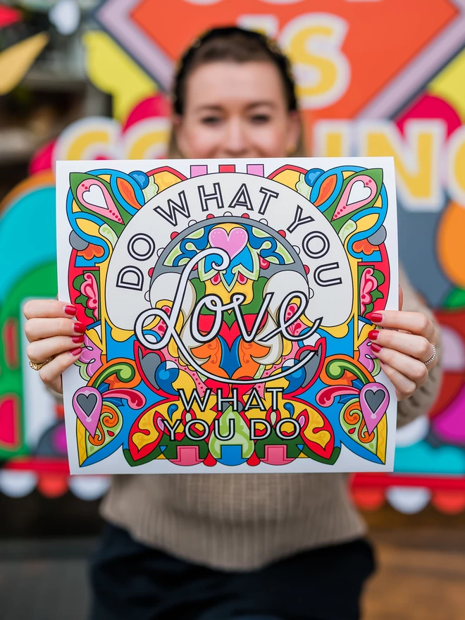A caucasian woman with brown hair holds in front of her the print which has “Do what you love, love what you do” written over a vibrant, rainbow coloured illustration.
