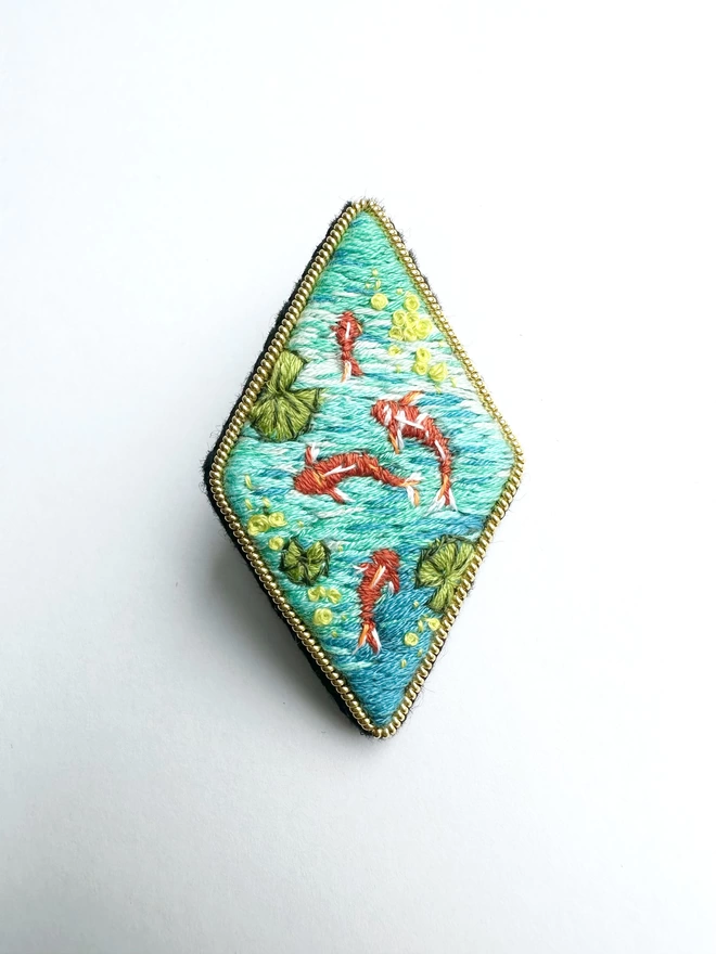 Diamond shaped brooch with image of coi carp fish swimming in a pond with on a plain background 