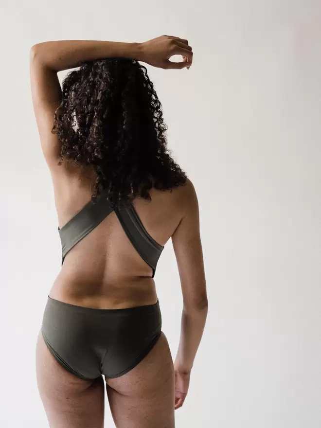 Back view of woman with long curly brown hair and one arm raised in studio wearing an olive green Davy J Sustainable Waterwear cutout swimsuit with wide cross back straps
