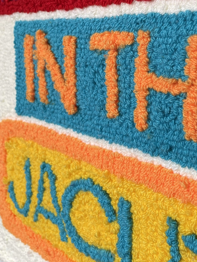 "in" & "the" in orange punch needle art a blue block with blue writing yellow & orange wool
