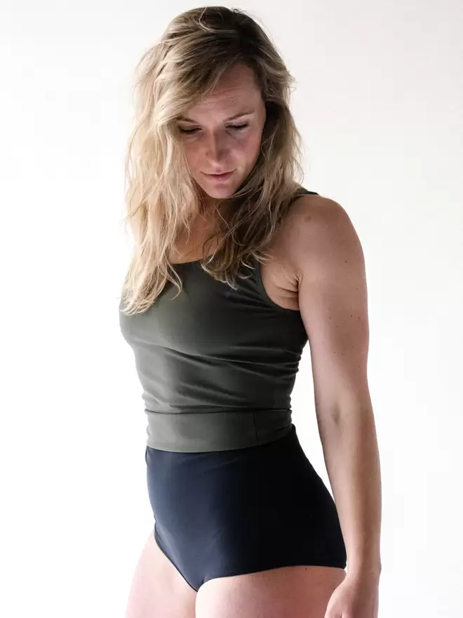 Blonde woman in studio looking down wearing Davy J Sustainable Waterwear olive green crop swim top and black high waist bikini briefs rolled down to show olive green lining