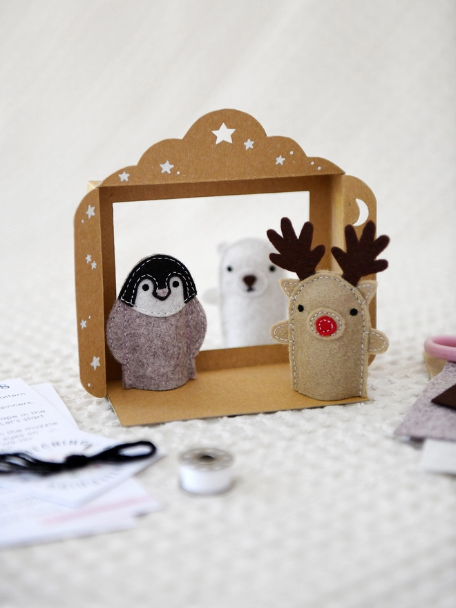 Three felt finger puppets, a reindeer, a penguin, and a polar bear, stand inside a small cardboard puppet theatre on an ivory fabric surface.